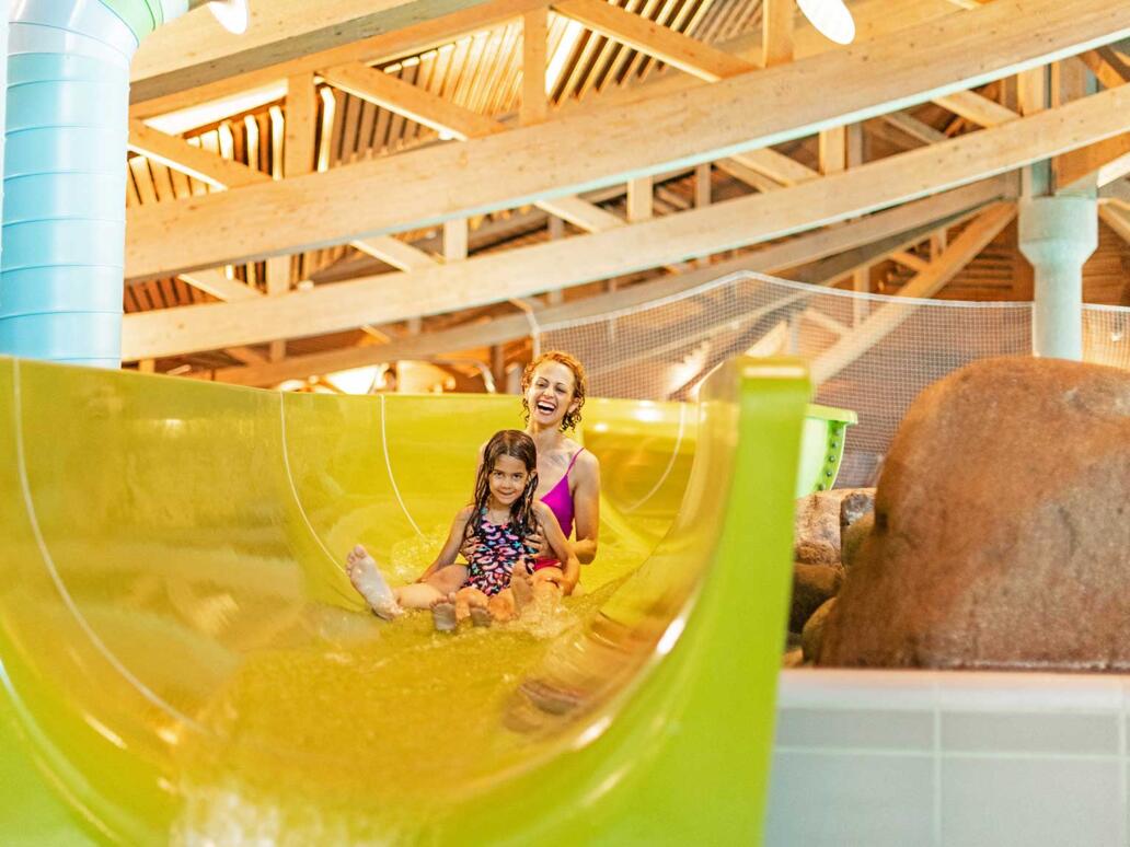 Zwei Kinder rutschen auf einer gelben Schlangenrutsche in der Kinderbadewelt eines Hallenbads ins Wasser.