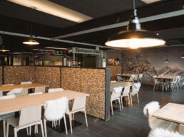 Gemütlicher Restaurantbereich mit Holztischen, weißen Stühlen, Fellauflagen und Holztrennwänden.