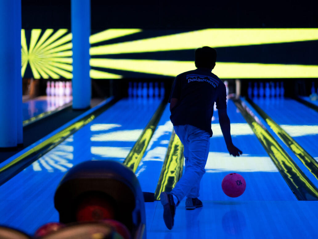 Person rollt eine pinke Bowlingkugel auf einer neonbeleuchteten Bowlingbahn im Hyper-Bowling-Bereich des Säntisparks.