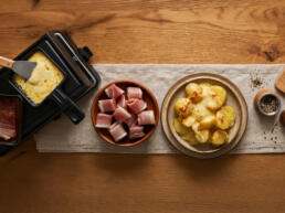 Raclette mit Kartoffeln, Speck und Käse auf einem Holztisch, von oben fotografiert.