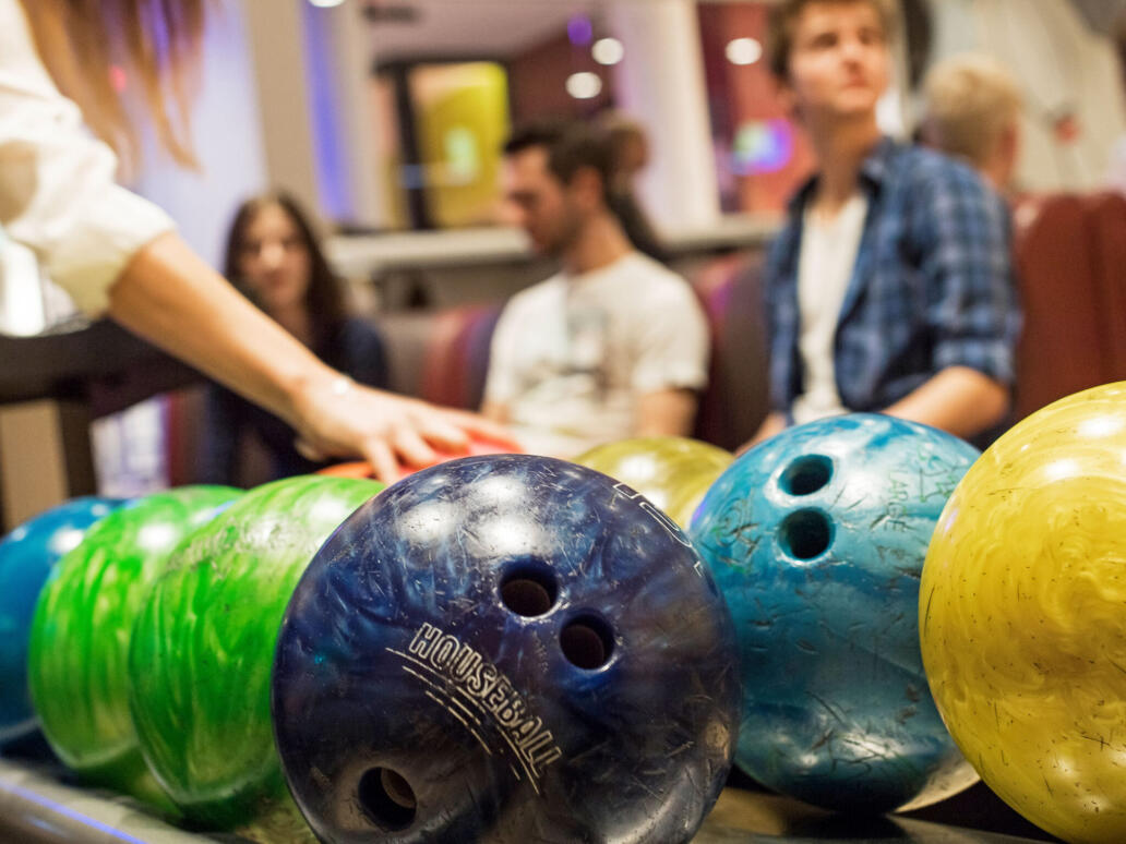 Mehrere farbige Bowlingkugeln liegen auf einer Kugelrückgabe bereit, während Personen im Hintergrund an einer Bowlingbahn stehen.